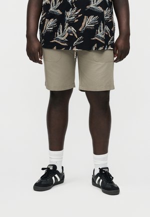 Asmuo dėvi smėlio spalvos šortus, juodus ir baltus sportinius batelius, baltas kojines bei juodą marškinėlį su smėlio spalvos ir baltų lapų raštais, stovintis prieš balto fono foną.