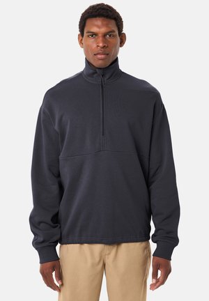 Mann trägt einen schwarzen Pullover mit Reißverschluss bis zur Brust und beige Hose, steht mit entspannten Armen vor einem einfarbigen Hintergrund.