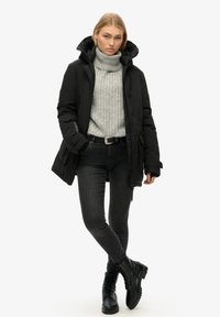 Zwarte gewatteerde parka met een hoge kraag, grijze geribbelde trui, donkere spijkerbroek en zwarte veterschoenen. Eenvoudige hardware-accenten en textuurstof.