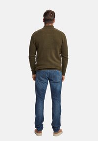 Uomo di spalle che indossa un maglione verde oliva, jeans blu e scarpe beige, in piedi contro uno sfondo chiaro e uniforme.