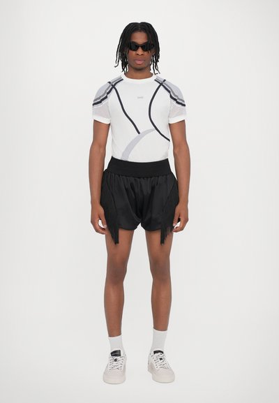 Kurze weiße Bluse mit grauen Akzenten und Netzärmeln, kombiniert mit schwarzen Shorts mit Fransen-Details. Sportliche weiße Sneaker und Socken.