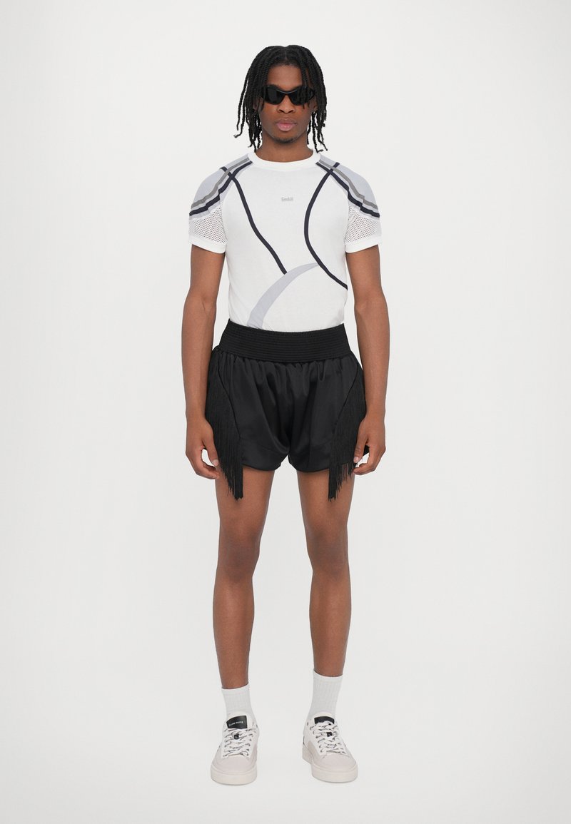 Kortærmet hvid top med grå detaljer og mesh-ærmer, parret med sorte shorts med frynse-detaljer. Sporty hvide sneakers og sokker.