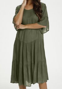Mujer vestida con un vestido suelto, de color verde oliva, a la altura de la rodilla, con pliegues en capas y mangas hasta los codos, de pie con los brazos cruzados.