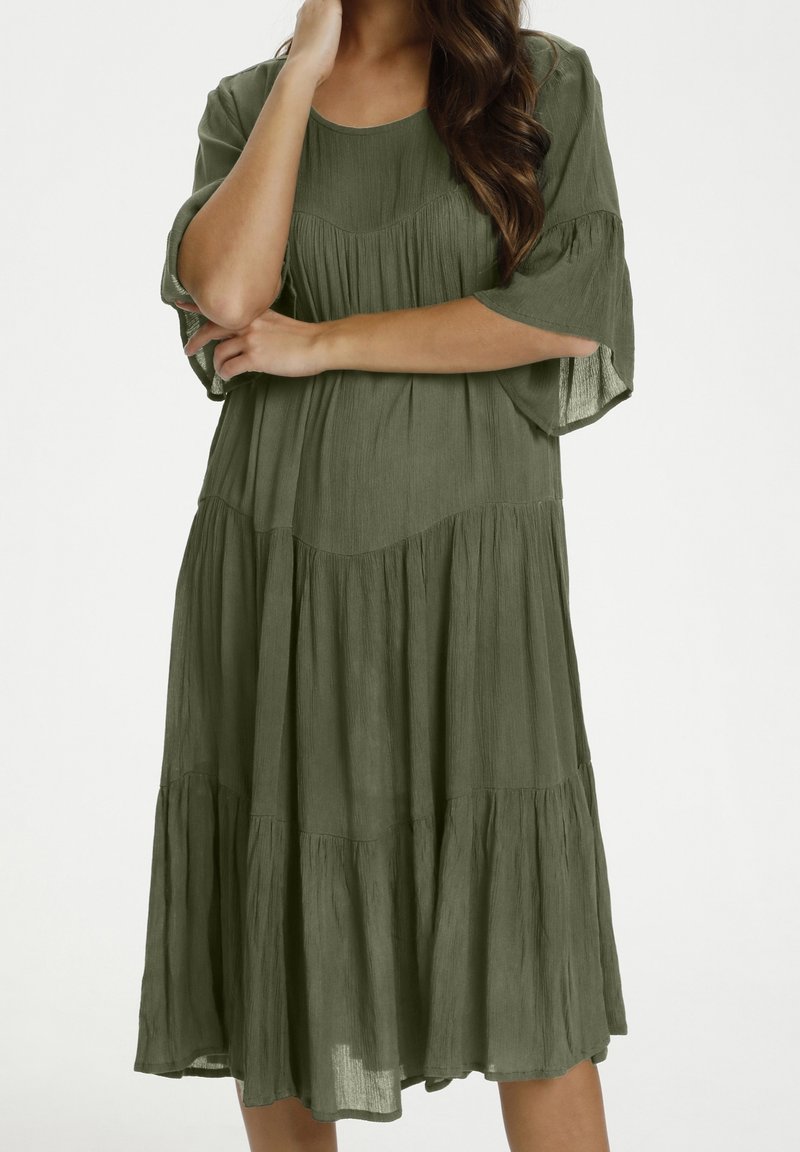 Mujer vestida con un vestido suelto, de color verde oliva, a la altura de la rodilla, con pliegues en capas y mangas hasta los codos, de pie con los brazos cruzados.