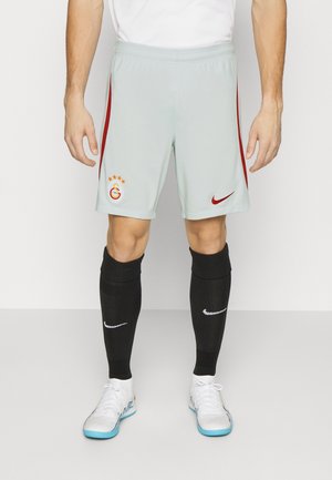 Athlète portant un short de sport gris clair avec des bandes rouges, des chaussettes noires montantes avec des logos blancs et des chaussures de football en salle blanches avec des semelles bleues.