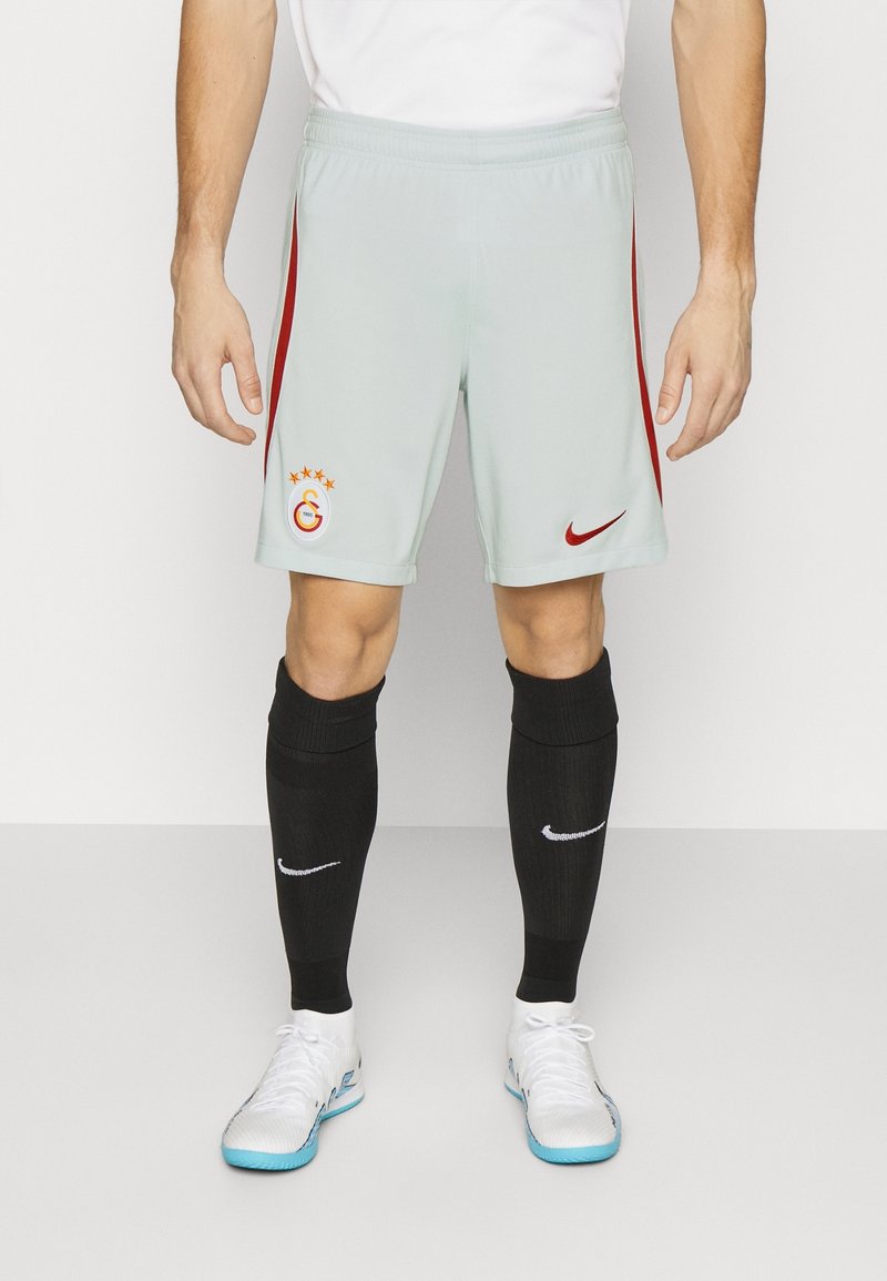 Athlète portant un short de sport gris clair avec des bandes rouges, des chaussettes noires montantes avec des logos blancs et des chaussures de football en salle blanches avec des semelles bleues.
