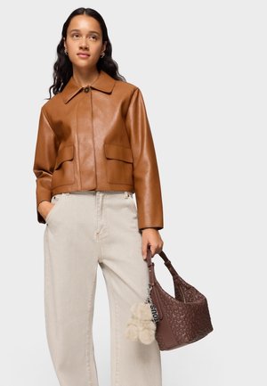 Femme portant une veste en cuir marron et un pantalon beige, tenant un sac à main marron tressé avec un porte-clés en fourrure.