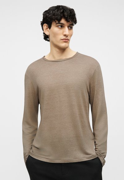 Νεαρός άνδρας με σγουρά σκούρα μαλλιά, που φοράει ένα ανοιχτό καφέ πουκάμισο με μακριά μανίκια και μαύρο παντελόνι, κοιτάζει προς τα αριστερά με τα χέρια στις τσέπες.