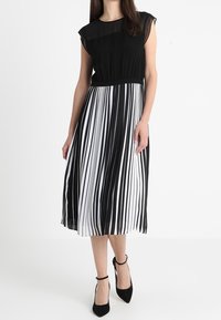 Schwarz-weiß gestreiftes Kleid mit tailliertem Oberteil, transparenten kurzen Ärmeln und knielangem, plissiertem Rock. Akzentuiert mit schwarzen High Heels.