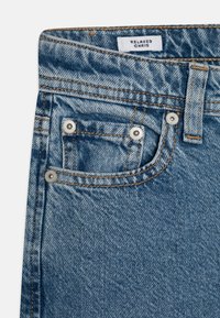 Shorts de denim en azul claro, con un corte holgado, costuras naranjas en contraste, remaches plateados y un pequeño bolsillo delantero. Etiqueta de marca visible.