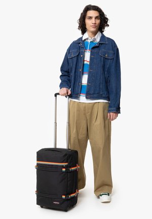 Black wheeled suitcase with rainbow strap accents, rectangular shape, durable fabric, and a retractable handle, positioned next to a person.