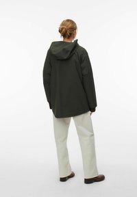 Personne aux cheveux blonds attachés en chignon, portant une veste à capuche vert foncé, un pantalon beige clair et des bottines marron, debout de dos.