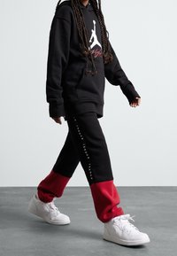 Czarna bluza z kapturem z białym logo Jordana, w połączeniu z czarno-czerwonymi spodniami dresowymi z bocznym logo. Dopełnieniem stroju są białe sneakersy.