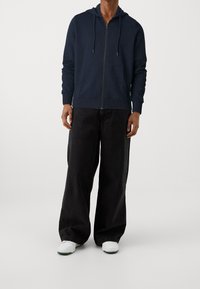 Felpa con zip di colore navy, dotata di due tasche frontali, cappuccio con coulisse e polsini a coste, abbinata a pantaloni larghi neri e sneakers bianche.