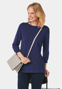 Dunkelblauer Pullover mit Dreiviertelärmeln, Rundhalsausschnitt und weicher Textur. Kombiniert mit einer hellgrauen, gesteppten Umhängetasche.