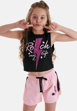 Junges Mädchen mit langen Haaren, das ein schwarzes ärmelloses Oberteil und pinke Shorts trägt, zeigt mit den Fingern auf ihre Wangen und macht ein verspieltes Gesicht.
