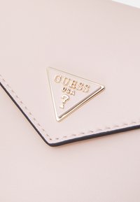 Blassrosa synthetische Handtasche mit einer dreieckigen goldfarbenen Logo-Plakette, versehen mit abgesteppten Kanten und einer glatten Textur.