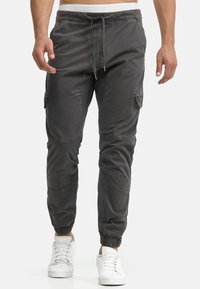 Dunkelgraue Herren-Cargohosen mit elastischen Bündchen, Kordelzugbund, Seitentaschen und Kniefalten. Getragen mit weißen Sneakern.