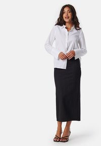 Chemise blanche à boutons avec des manches longues, associée à une jupe midi noires à rayures. Modèle portant des sandales noires à lanières.