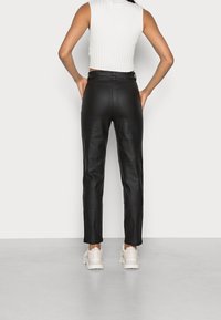 Pantaloni in finta pelle nera con vestibilità slim, caratterizzati da una vita media, due tasche posteriori e una texture liscia. Indossati con un top bianco senza maniche.