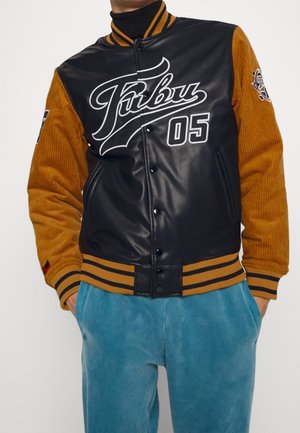 Sort og brun varsity-jakke med teksten "Fubu 05", ribstrikkede, stribede manchetter og krave, kombineret med blå bukser, hænderne i lommerne.
