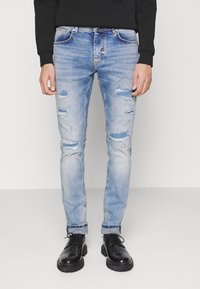 Jeans de denim desgastados de color azul claro con varios desgarros y un ajuste slim, combinados con botas negras con cordones y una camiseta de manga larga negra.