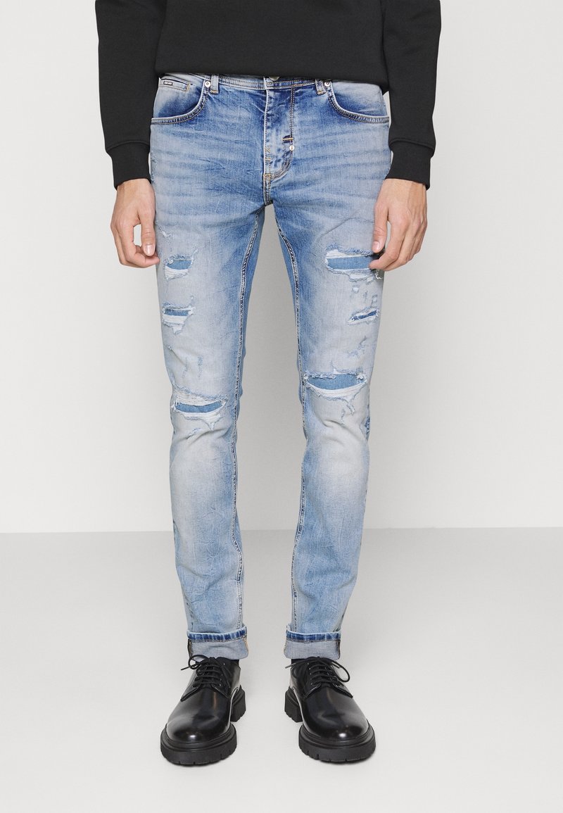 Jeans de denim desgastados de color azul claro con varios desgarros y un ajuste slim, combinados con botas negras con cordones y una camiseta de manga larga negra.