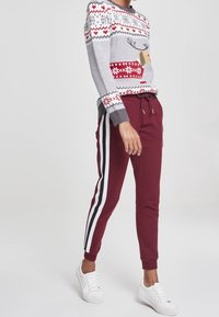 Grauer Pullover mit einem Rentierdesign, roten Akzenten und einem Schneeflockenmuster, kombiniert mit burgunderfarbenen Jogginghosen mit weißen Seitenstreifen und weißen Sneakern.