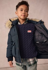 Suéter de punto trenzado en color navy debajo de una chaqueta acolchada verde oscura con capucha forrada de plumas; combinado con una camisa de cuadros y jeans azules.