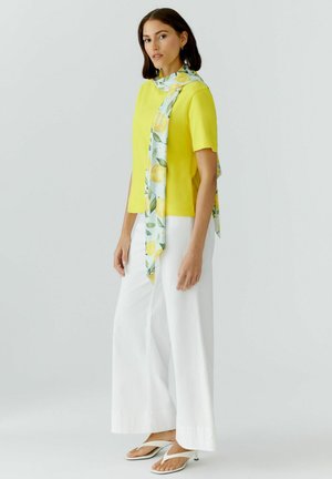 Femme debout, portant un haut jaune à manches courtes, un pantalon blanc à jambes larges, des sandales blanches à talons et une écharpe à motif citron.