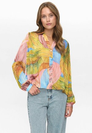 Kvinna med långt vågigt hår, iförd en färgglad knapp-up blus med abstrakta mönster och ljusblå jeans, stående mot en neutral bakgrund.