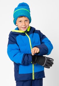 Blauwe en marineblauwe waterdichte jas met een groene rits en capuchon, zwarte handschoenen en een blauwe gebreide beanie met groene strepen en een pompon.