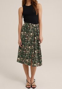 Černý tílko a zelená květinová midi sukně s předním rozparkem. Sukně má strukturovaný vzor s hnědými a krémovými akcenty. Černé sandály na pásky.