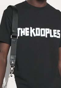 Svart t-shirt i bomull med fet vit text som säger "THE KOOPLES." Kort ärm och rundad halsdesign. Minimalistisk stil.