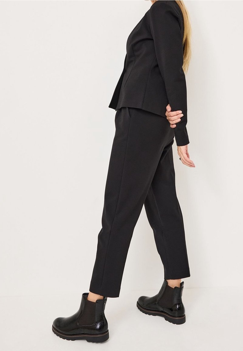 Femme portant un pantalon noir sur mesure, un blazer assorti et des bottines noires brillantes, sur un fond clair uni.