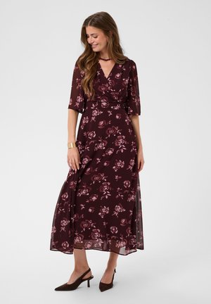 Femme portant une robe portefeuille fleurie violet foncé avec des manches jusqu'au coude, accessoirisée d'un bracelet, d'un collier et de talons slingback foncés.