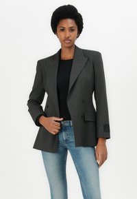Siv šivalni blazer z urejeno silhueto, dvorednim zapenjanjem in poševnimi žepi. V kombinaciji z oprijetimi jeans hlačami in črno majico.
