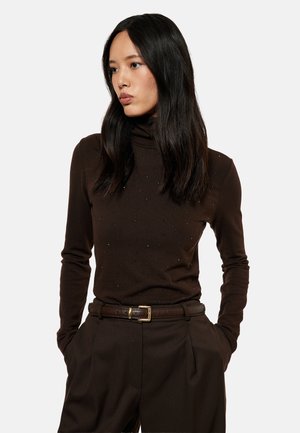Giovane donna con lunghi capelli neri che indossa un maglione dolcevita marrone scuro e pantaloni a vita alta abbinati con cintura, mani nelle tasche.