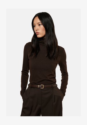 Giovane donna con lunghi capelli neri che indossa un maglione dolcevita marrone scuro e pantaloni a vita alta abbinati con cintura, mani nelle tasche.