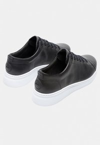 Svarta lädersneakers med en slät yta, kontrasterande vita gummisulor och sex hål för snörning. Baksidan har en cirkulär detalj.