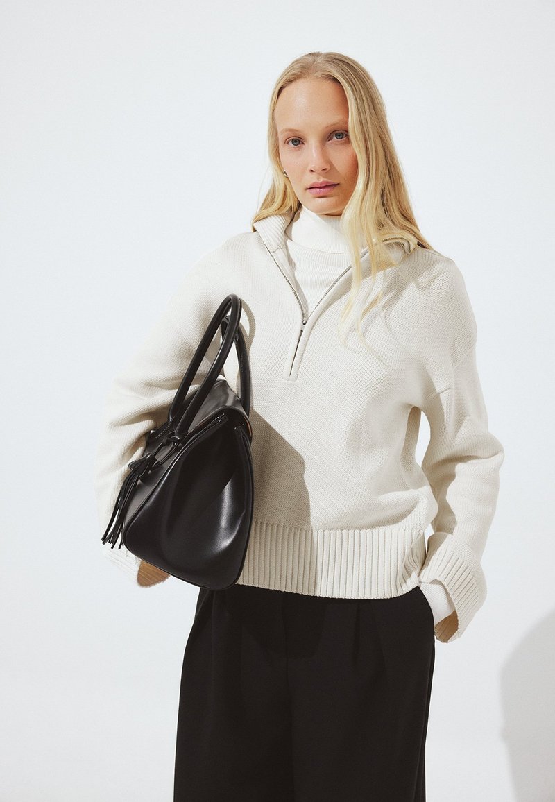 Frau mit langen blonden Haaren, die einen cremefarbenen Pullover mit halbem Reißverschluss und schwarze Hose trägt, hält eine große schwarze Ledertasche über ihre Schulter.