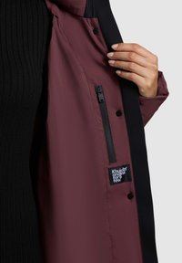 Tumepruun jakk mustade detailidega, millel on tõmblukuga tasku, trukid ja sees kaubamärgi silt. Materjal näib kerge ja sile.