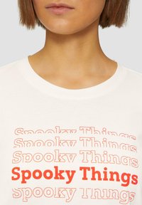 Witte katoenen t-shirt met gestapelde tekst "Spooky Things" in verschillende lettertypen, waarbij de grootste in felrood is en de kleinere in oranje en zwart.
