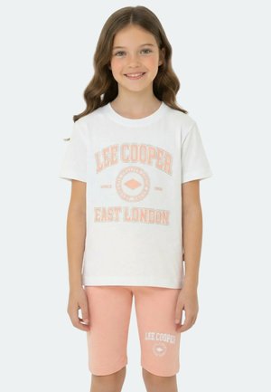 Fille souriante aux longs cheveux porte un T-shirt blanc Lee Cooper et un short pêche à hauteur des genoux avec le logo Lee Cooper sur la jambe du short.
