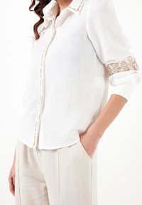 Camicia bianca con bottoni realizzata in tessuto leggero. Presenta dettagli in pizzo floreale sulle maniche e una consistenza morbida. Indossata infilata in pantaloni chiari.