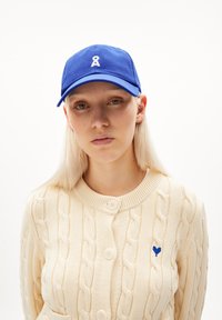 Blauwe katoenen baseballcap met een wit logo, gecombineerd met een crèmekleurige kabelbrei cardigan met grote knopen en een klein blauw hartdetail.