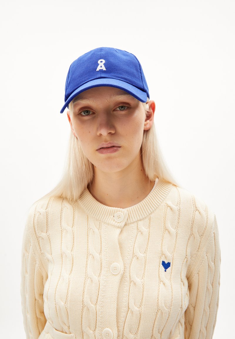 Blauwe katoenen baseballcap met een wit logo, gecombineerd met een crèmekleurige kabelbrei cardigan met grote knopen en een klein blauw hartdetail.