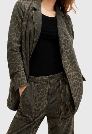 Personne portant une veste imprimé léopard vert olive et un pantalon assorti avec un haut noir, mains dans les poches, vue des épaules jusqu'au milieu des cuisses.