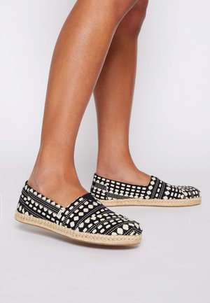 Jambes portant des espadrilles à enfiler à motifs noirs et blancs avec des semelles en jute tissé sur un fond clair uni.