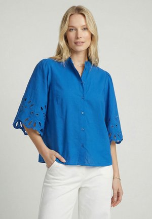 Femme portant un chemisier bleu vif à boutons avec des manches découpées brodées et un pantalon blanc, debout avec une main dans la poche.